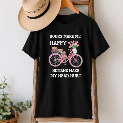May include: Black t-shirt with the text "BOOKS MAKE ME HAPPY HUMANS MAKE MY HEAD HURT". The graphic features a pink bicycle carrying books and flowers. A straw hat hangs above the shirt on a wooden rack.