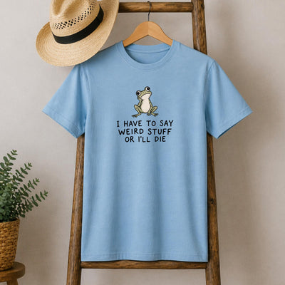 May include: A light blue short-sleeved t-shirt featuring a graphic of a small green frog with large eyes. Below the frog, the text reads: I HAVE TO SAY WEIRD STUFF OR I'LL DIE. The shirt has a crew neckline and is displayed on a wooden hanger.