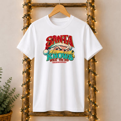 May include: A white short-sleeved t-shirt featuring a graphic of Santa Claus peering over a yellow newspaper. The text reads SANTA KNOWS WHAT YOU DID and he has receipts in red, teal, and black lettering across the chest area.
